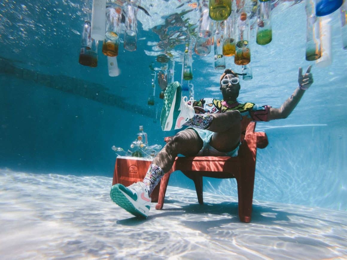 un bărbat este asezat pe un scaun de plaja sub apa, intr-o piscina, in timp ce in jurul lui plutesc mai multe sticle și pahare