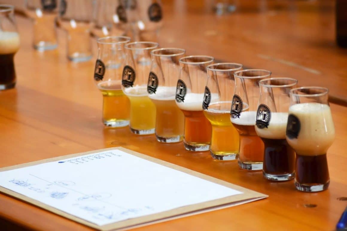 mai multe pahare de bere, umplute pe jumătate cu diferite tipuri de bere sunt asezate pe o masă cu un clipboard în fața lor.