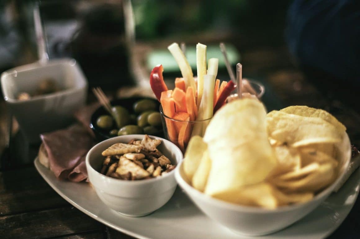 un platou de aperitivo, cu masline, biscuiti s arati, chipsuri si legume crude, asezate pe un platou alb pus pe o masă de lemn