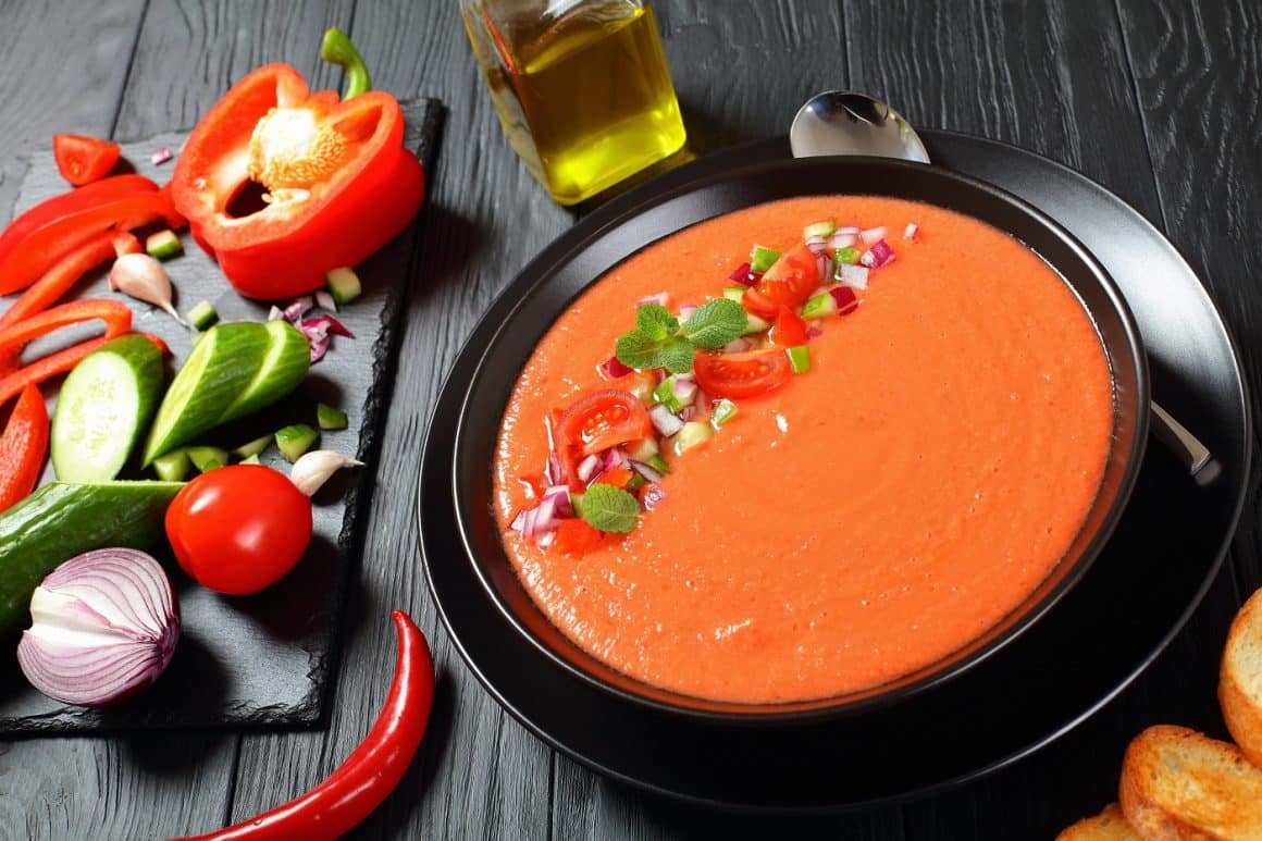 portion of delicious gazpacho in black bowl - spanish style cold summer tomato soup, traditional recipe. ingredients on wooden table, horizontal view from above, close-up