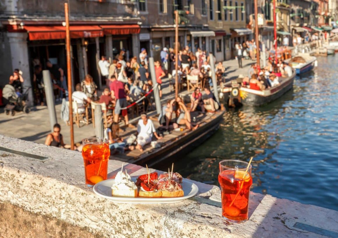 doua pahare cu aperitivo italian aperol spritz și un platou cu bruschete sunt asezate pe balustrada de piatra a unui pod din venetia, iar in fundal se vad mai multe barci incarcate cu oameni, pe un canal