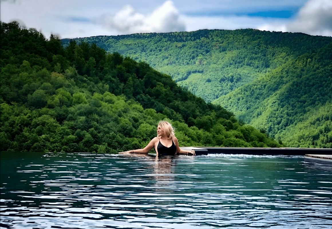 femeie blonda se relaxeaza în piscină la munte, iar in fundal dealurile pline de verdeață
