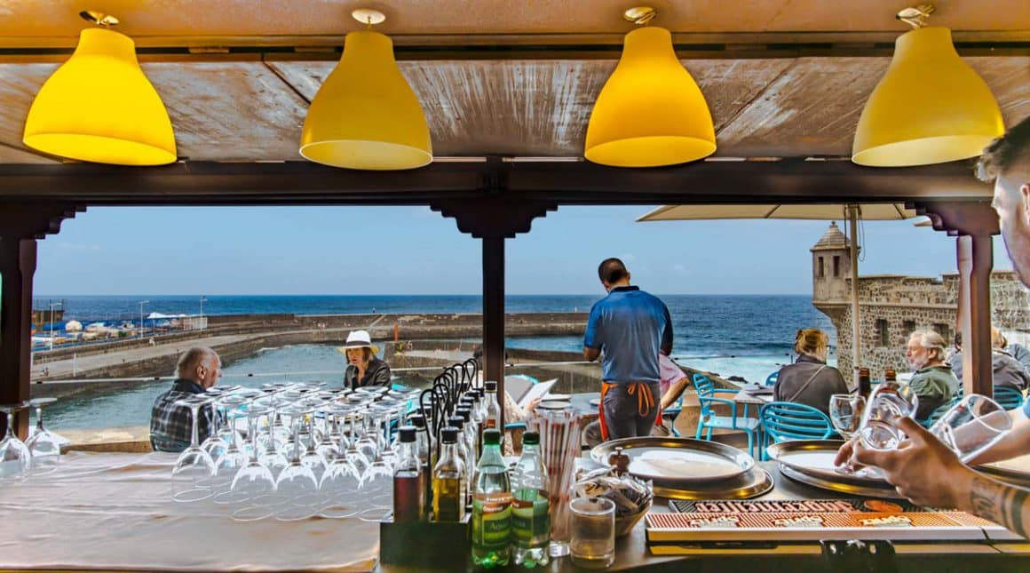 mai multi oameni iau masa pe terasa restaurantului Cofradía de Pescadores de Puerto de la Cruz, in fundal se vede marea iar in primul plan sunt patru corpuri de iluminat galbene