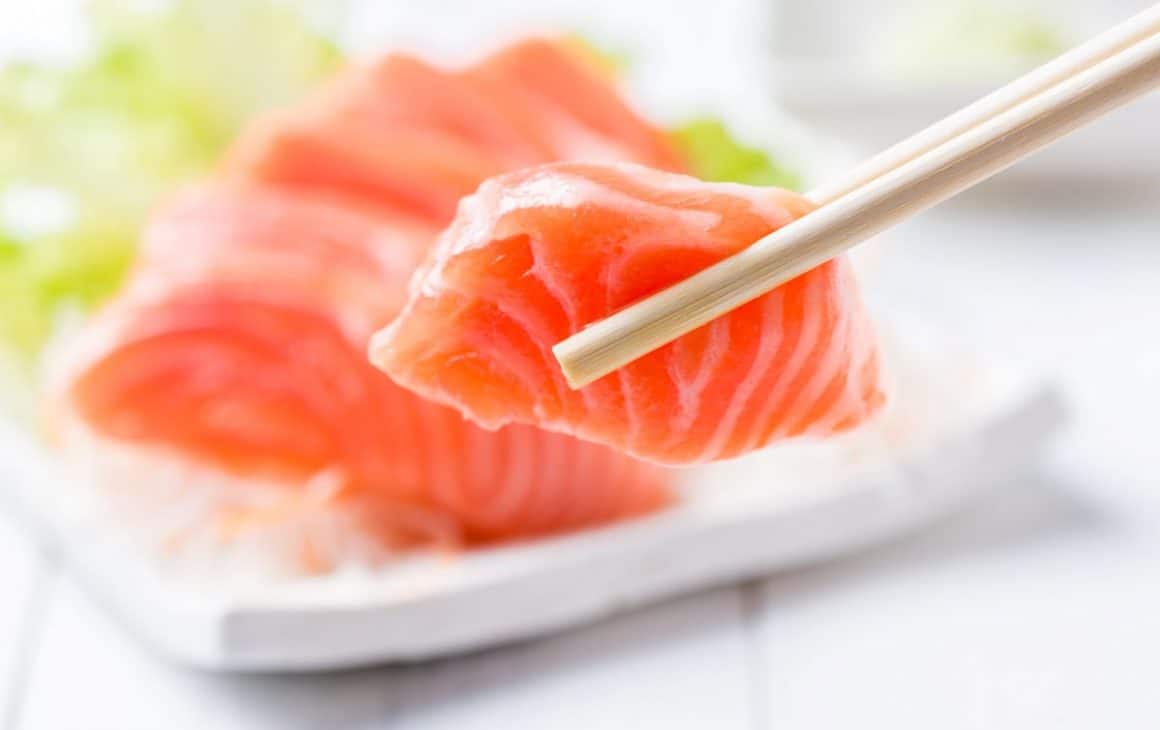 mai multe bucati de peste sashimi sunt asezate pe un vas ceramic alb, iar una dintre ele este ridicata cu ajutorul betisoalrelor chinezesti
