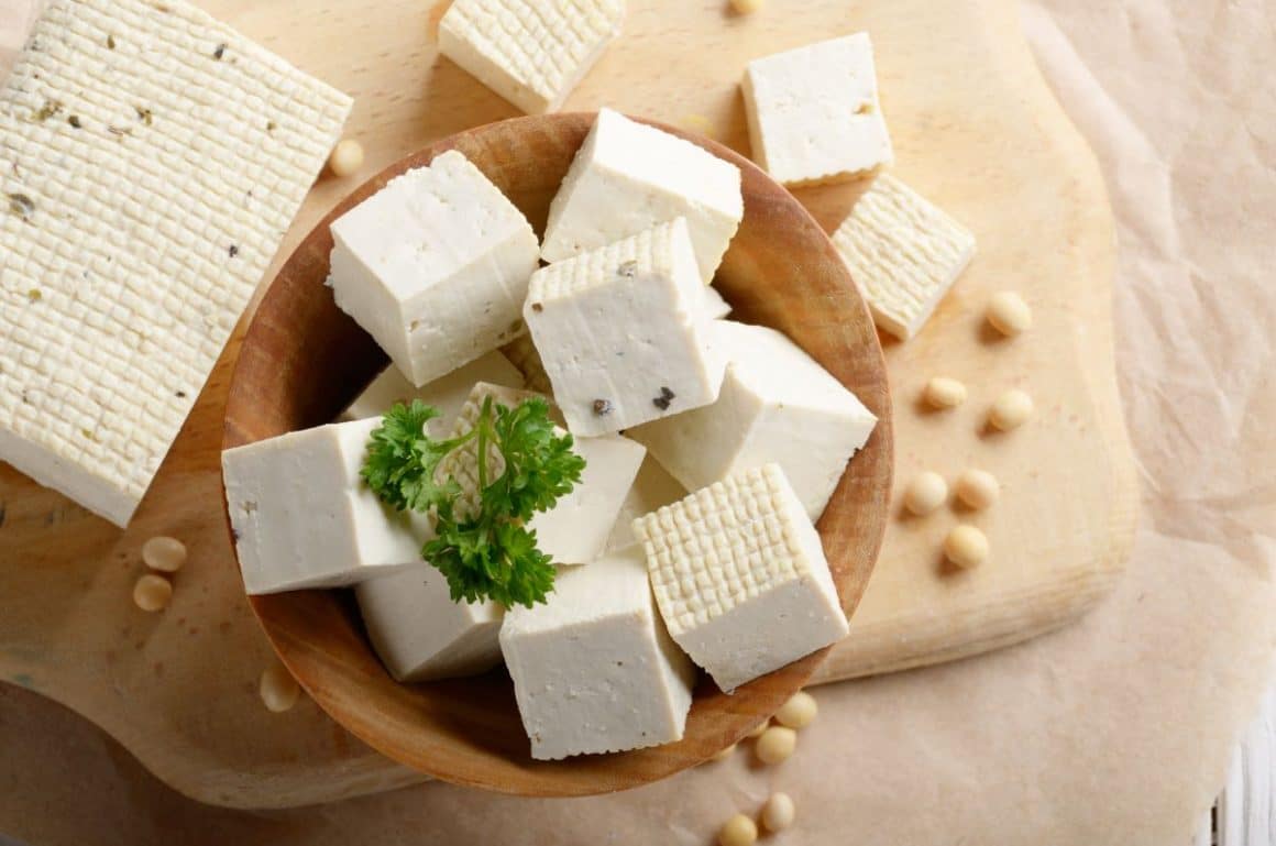 mai multe bucatele de tofu sunt asezate intr-un bol de lemn, pus pe omasa alaturi de o bucata mai mare de tofu