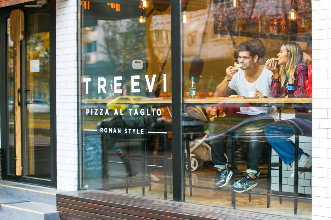 Cuplu care mananca pizza asezati la o masa inalta, de bar, in fata unei ferestre stradale la Treevi, fotografiați din exterior