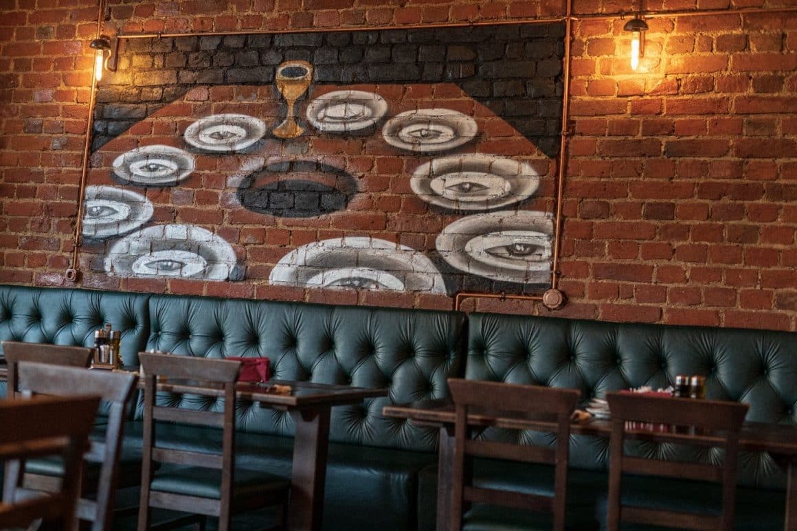 detaliu din restaurantul Costelaria - mese si o canapida lunga din piele verde, iar in spate un perete din caramida, pe care sunt pictate farfurii, 