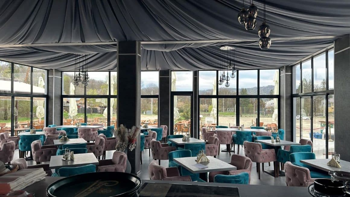 mai multe fotoliii bleu și roz sunt asezate langa mese, intr-o sală de restaurant cu geamuri mari și vualuri  textile pe tavan