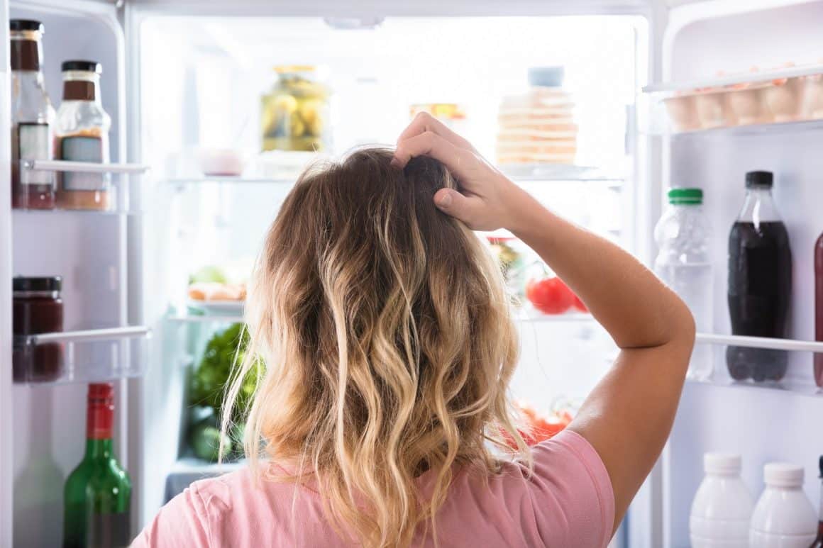 Femeie fotografiata din spate, se uita confuza în frigider. trucuri pentru cum să depozitezi mâncarea