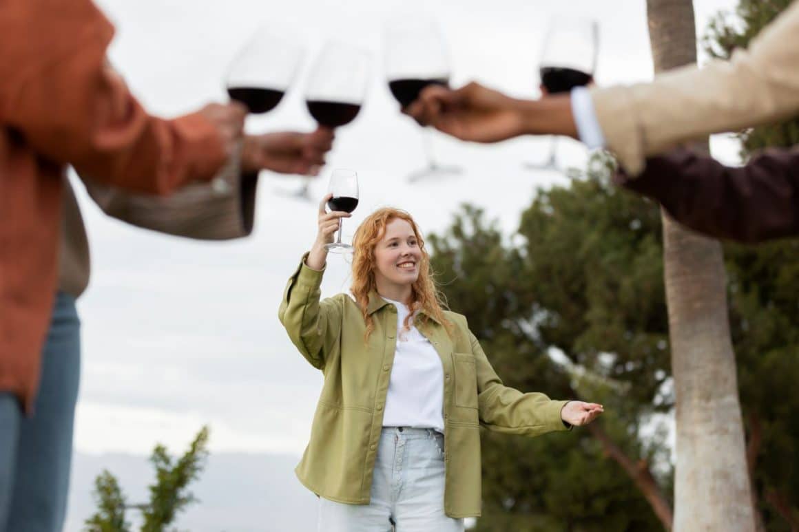 o femeie tânără ține în mână un pahar de vin ecologic și toastează cu alte trei persoane cu pahare de vin în mână. Ce sunt vinurile ecologice