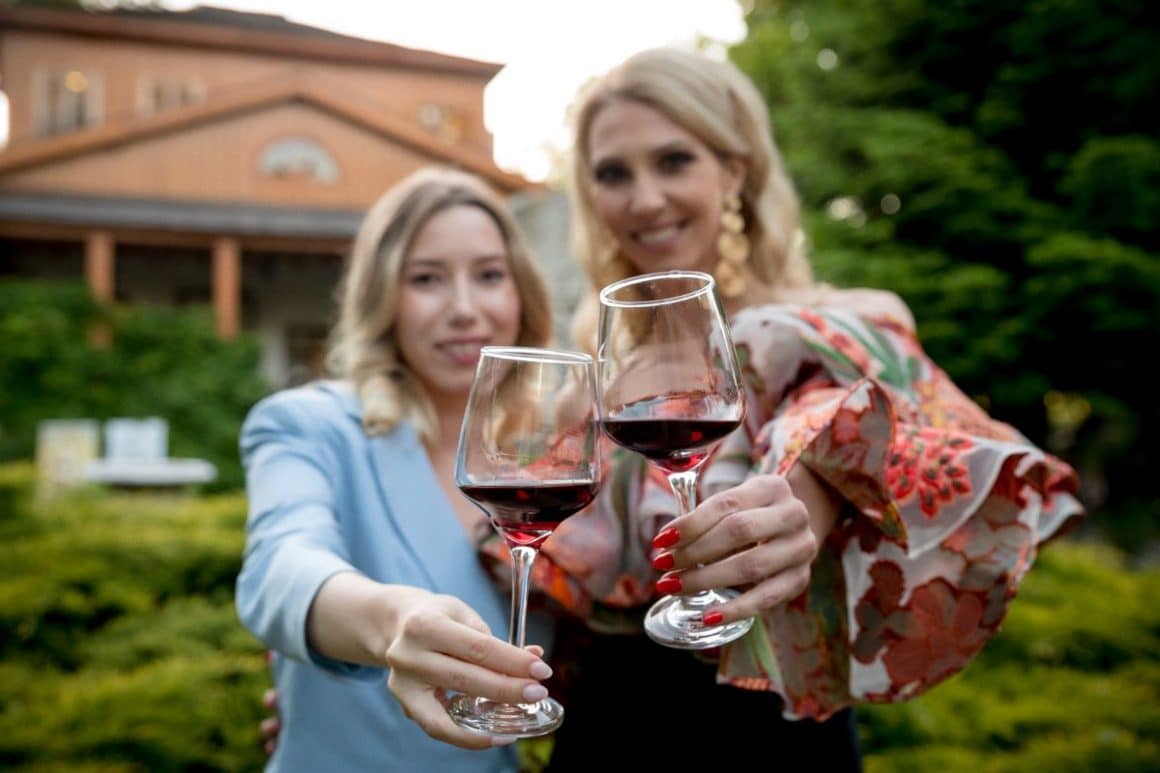 două femei toasteaza cu pahare de vin in mana, in gradina unei crame de pe drumul vinului