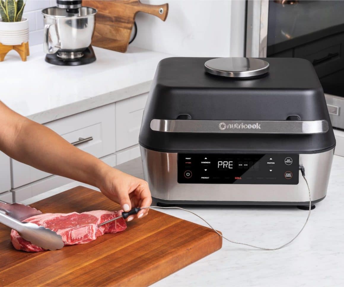 o mana de barbat înfige un termometru de carne într-un steak de vită, asezat pe un tocător de lemn, in fata unui gadgeturi de bucatarie
