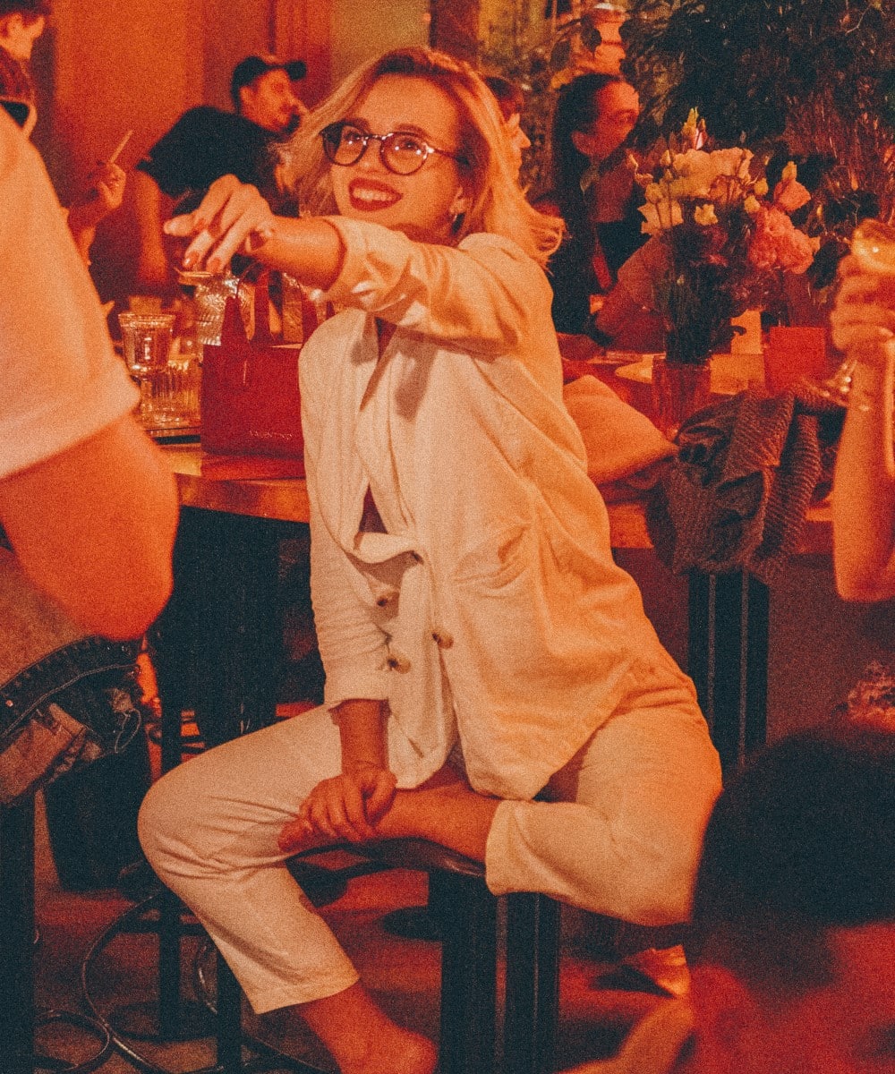 femeie tanara, blonda,, cu ochelari, in costum alb, sta pe un scaun de bar si intinde razand un pahar de cocktail