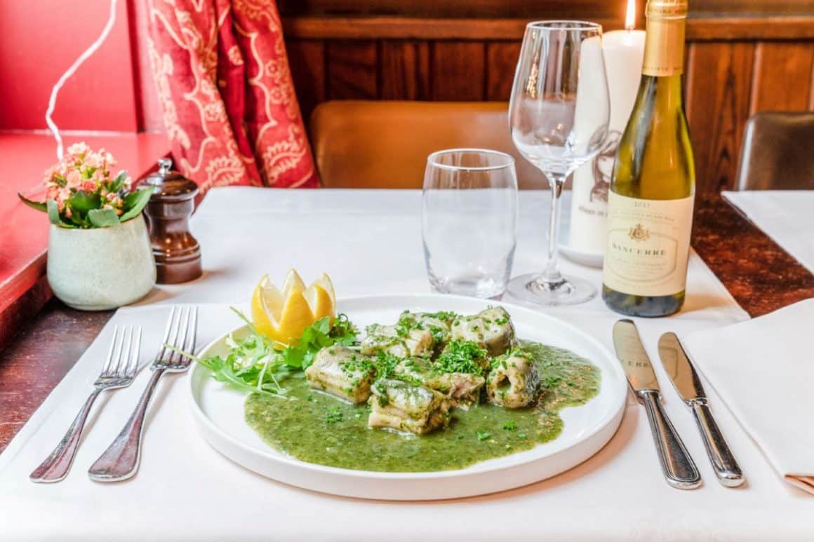 o portie de anghilă în sos verde, un fel tipic de mâncare în Bruges, cu tacâmuri și o sticlă de vin alb. De mâncat în Bruges