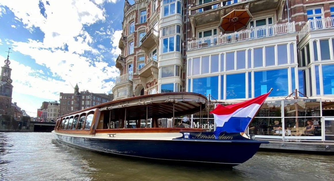 o barcă pentru croaziere turistice, cu steagul olandei arborat în vârf, trece prin fata uni restaurant, pe un canal din amsterdam