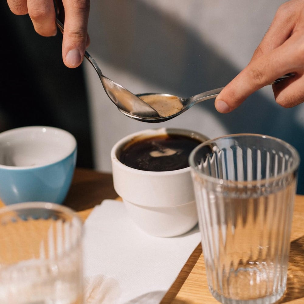 un barista ridică caimacul unei cafele dintr-o cească, cu ajutorul a două lingurițe