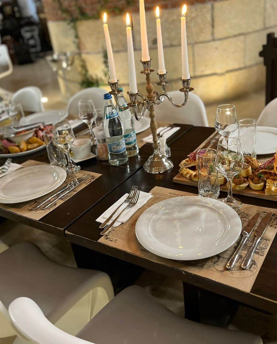 o masă de restaurant este asezata și pregtită să primească oaspeți, cu farfurii, tacâmuri și un candelabru pe ea