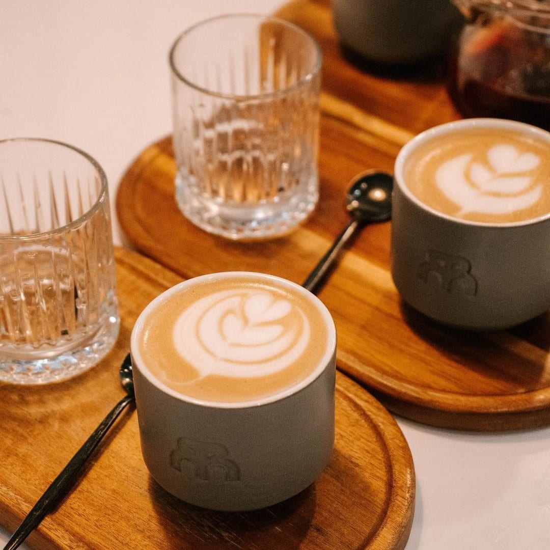 două cești de cafea cu lapte sunt asezate lângă două pahare de apă, pe două suporturi de lemn
