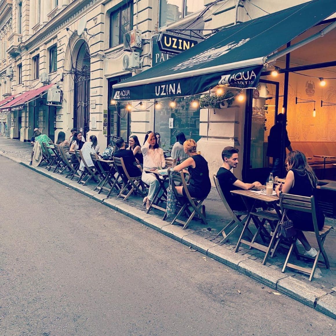 mai multi tineri socializează la mesele de la exterior, de pe trotoarun din fața unei cafenele micuțe din bucuresti