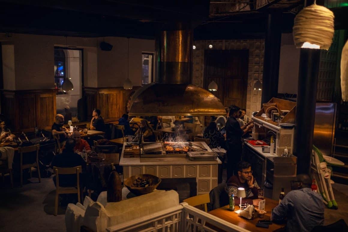Mai mulți oameni iau masa in restaurant City Grill, seara, luminat difuz, cu un cuptor mare in mijlocul incaperii. Restaurante românești București