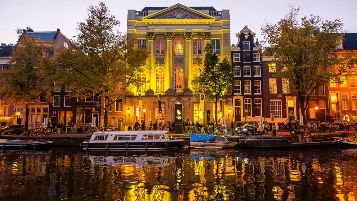 o clădire luminată de pe malul unui canal din amsterdam, pe care plutesc cțteva bărci
