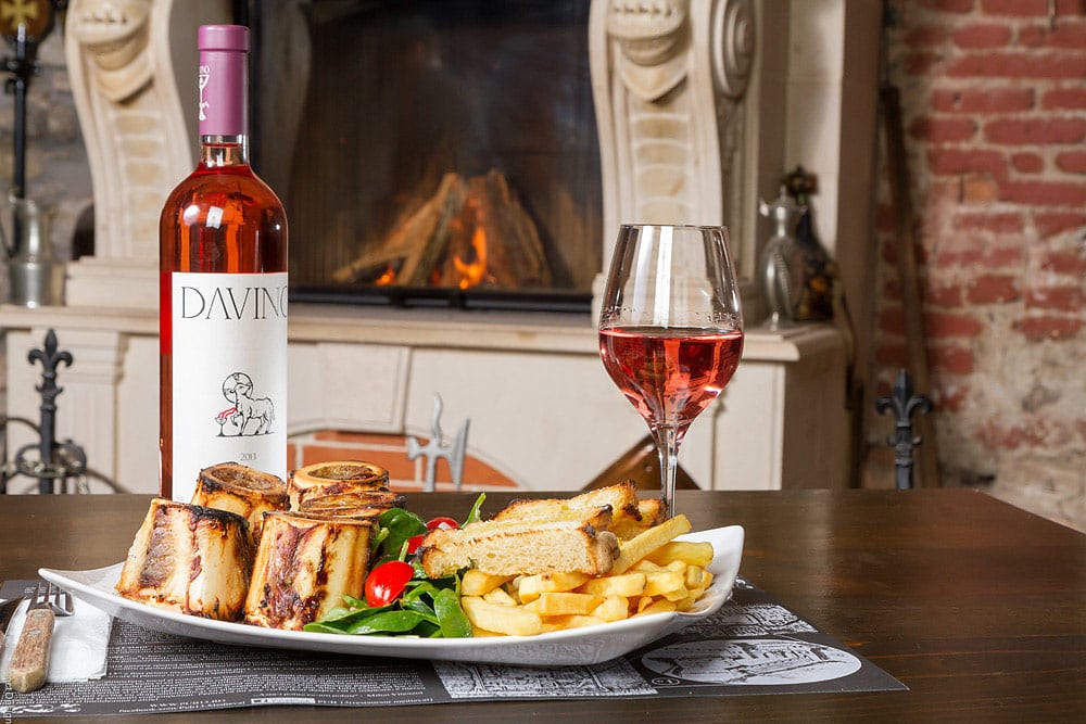 o portie generoasă de carne și cartofi prăjiți cu salată este asezata pe o masă de lemn, alături de o sticlă de vin și un pahar