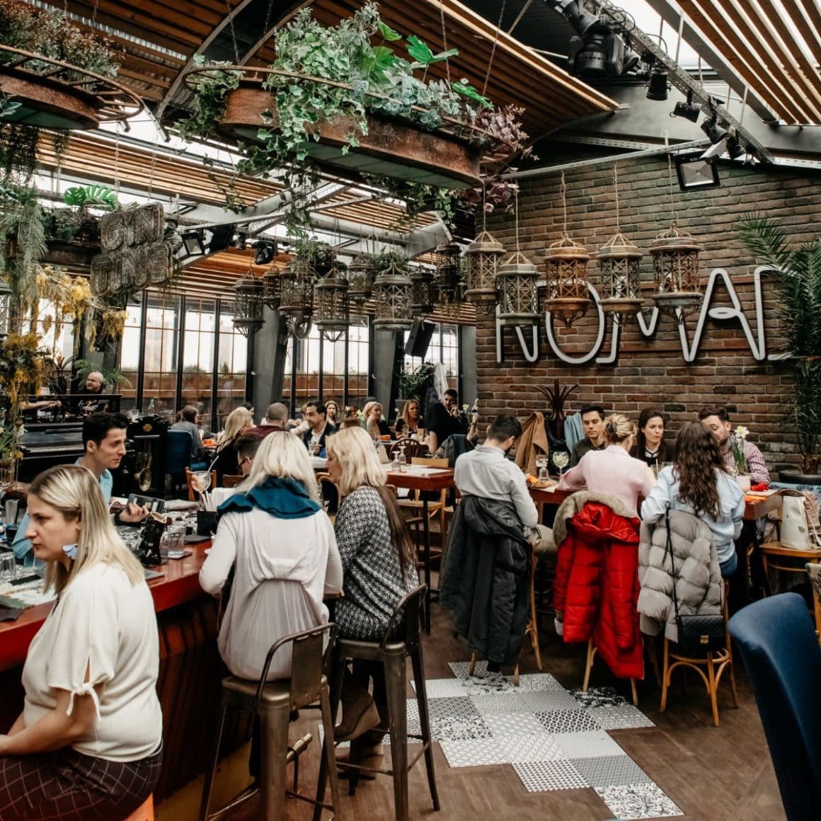 mai multi tineri socializează la mesele unui restaurant pentru grupuri mari din Bucuresti, cu multe plante curgătoare și riflaje din lemn