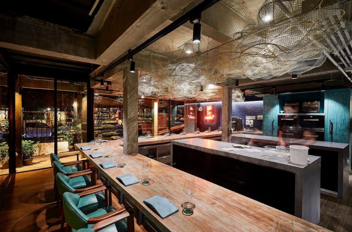 interiorul unui restaurant, cu mese lungi de lemn, și luminat discret