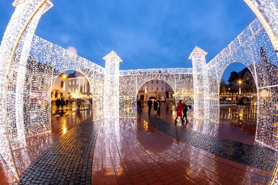 mai mulți oameni trec prin fața intrării in târgul de crăciun de la Timișoara, cu instalații luminoase