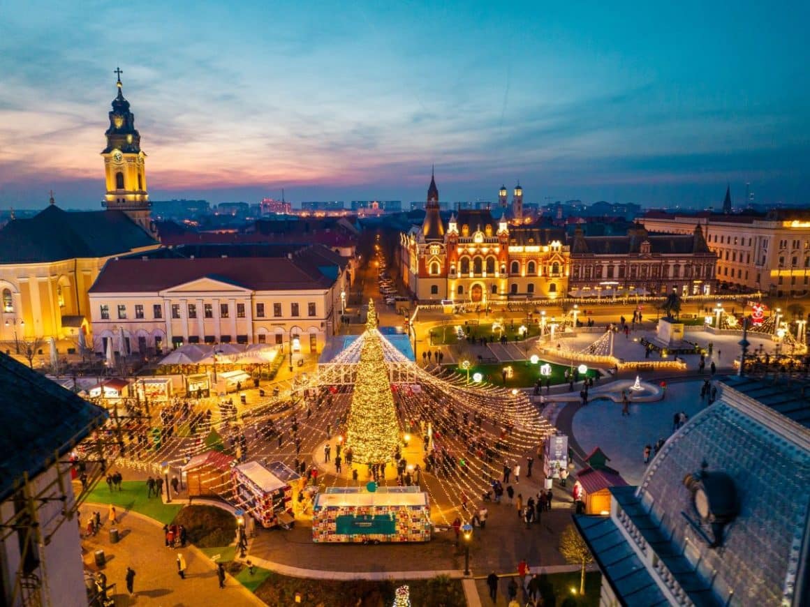 o fotografie aeriana cu casutele de la targul de craciun din oradea și cateva cladiri emblematice pentru oras, luminiate de beculetele. Unul din cele mai frumoase târguri de Crăciun din România 2023