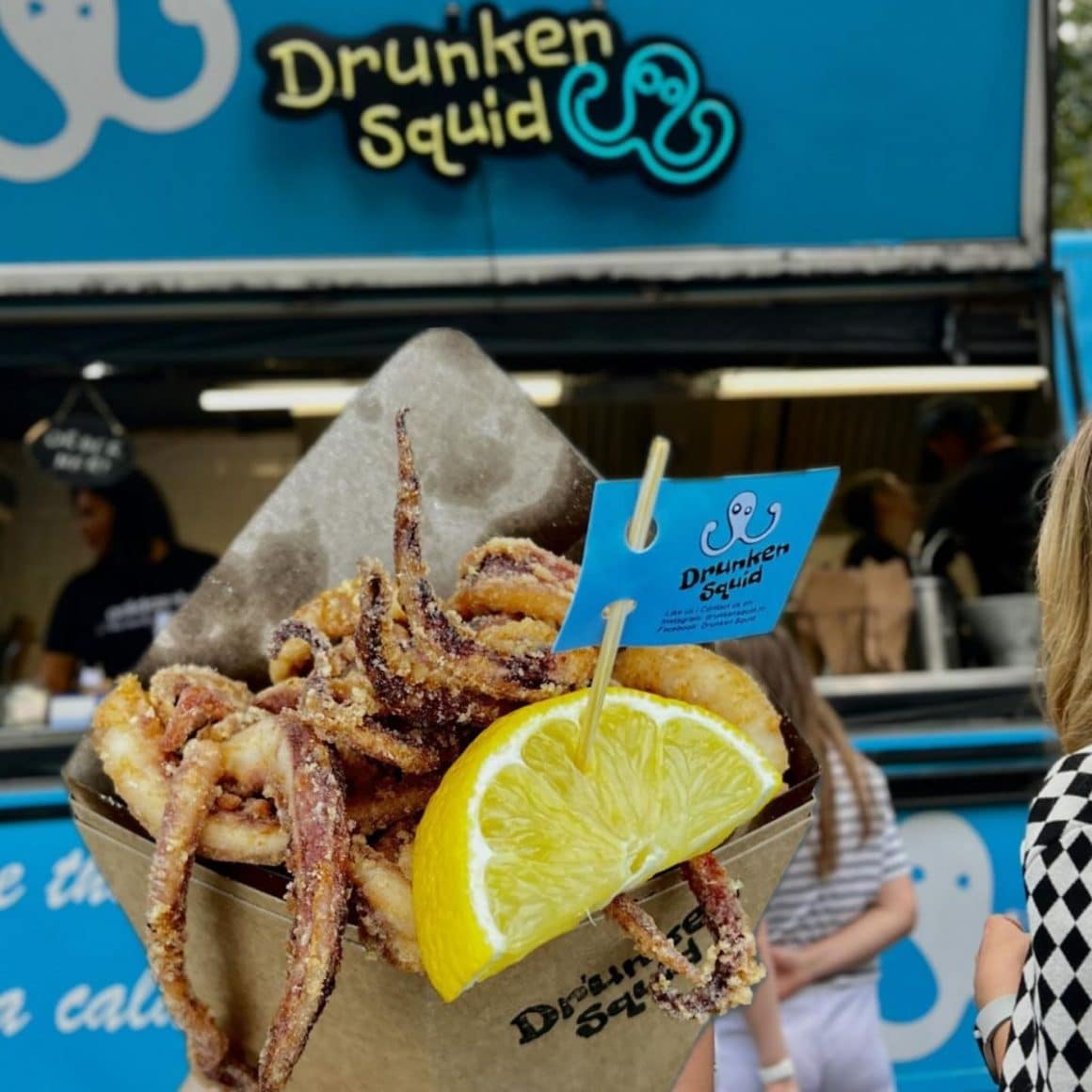 o femeie târnără ține în mână un recipient cu calamar pane în fața unui food truck la un festival. Cele mai bune foodtrucks