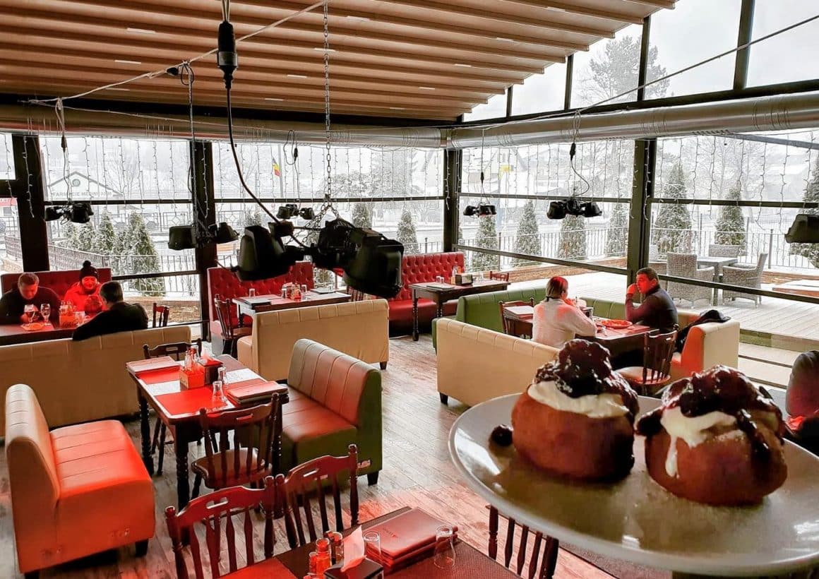 2 oameni asezati la mese intr-un resstaurant inchis cu pereti de sticla, prin care se vede zapada afara. restaurant Vatra Regală. Restaurante din Predeal