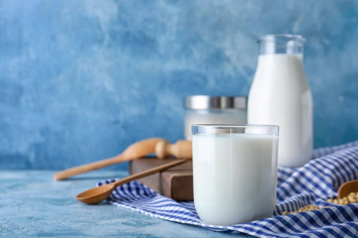 un pahar cu lapte, așezat pe un ștergar alb cu albastru, iar în fundal se vede o sticlă de lapte și câteva ustensile de bucătărie din lemn