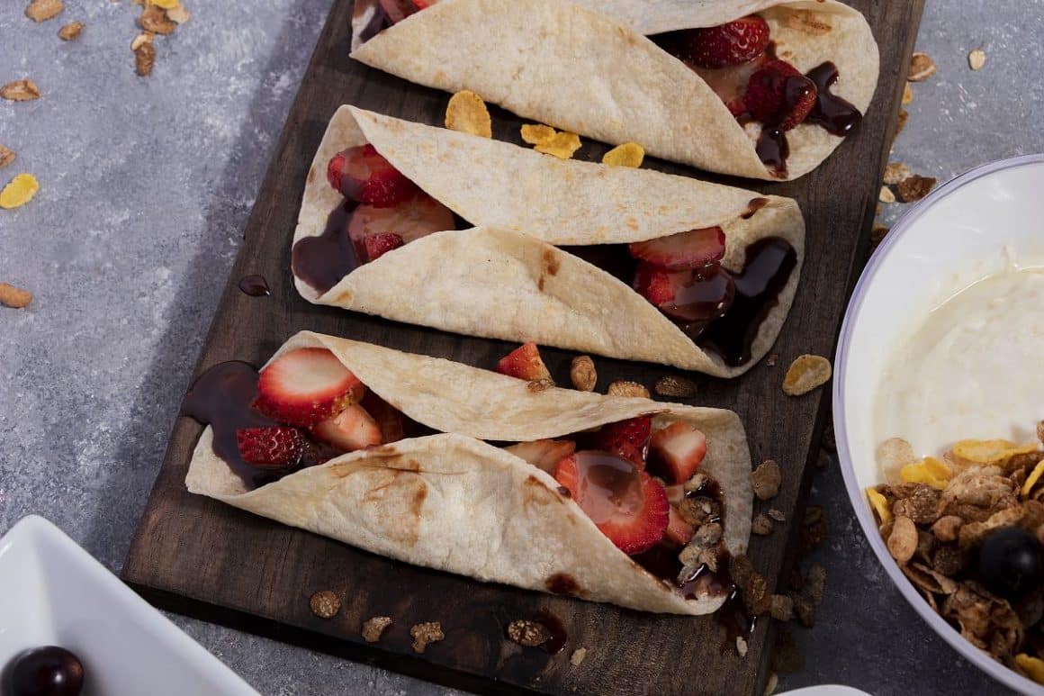 trei Strawberry burrito sunt asezate pe un tocător de bucătărie, cu căpșuni și sos de ciocolată