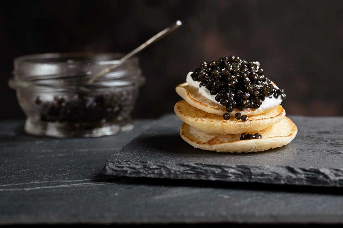 un recipient cu caviar este asezat lângă un stack de clătite peste care au fost puse niște icre negre. food trends 2024