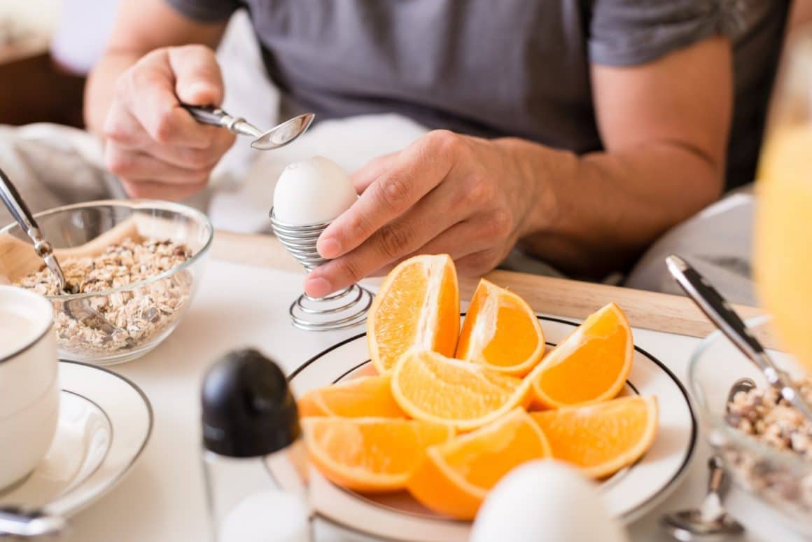 un barbat se pregateste sa sparga un ou fiert, pe care il serveste alaturi d eun bol de ovaz si portocala. Imagine concept pentru dieta cu oua fierte. Diete populare