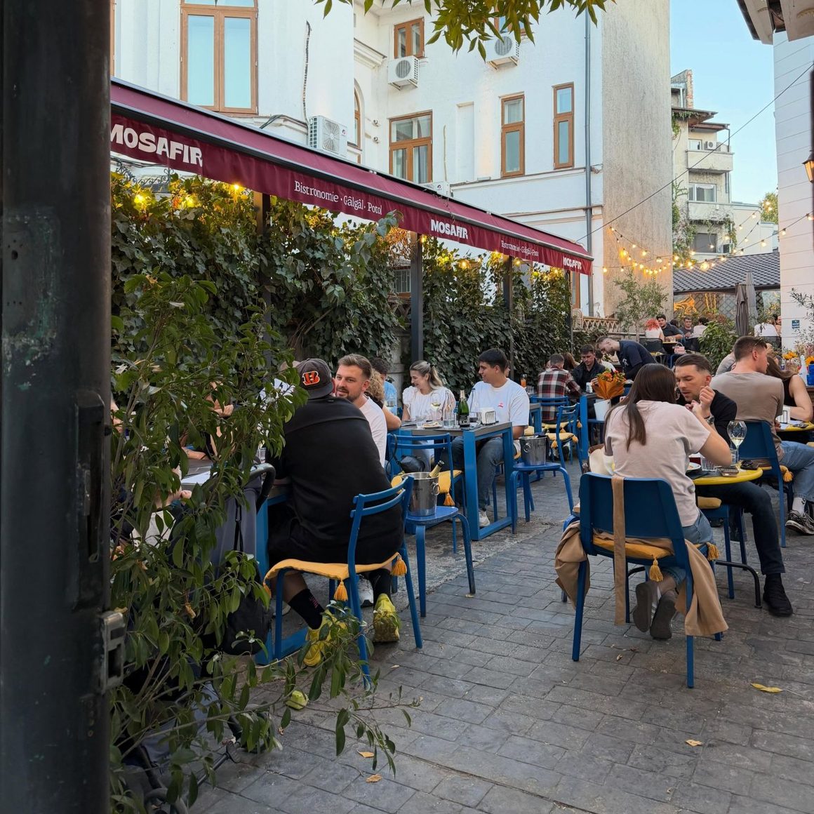 Bistrouri București: terasa Mosafir, cu mobilier albastru, vegetație și lumini ambientale, într-un spațiu urban animat.