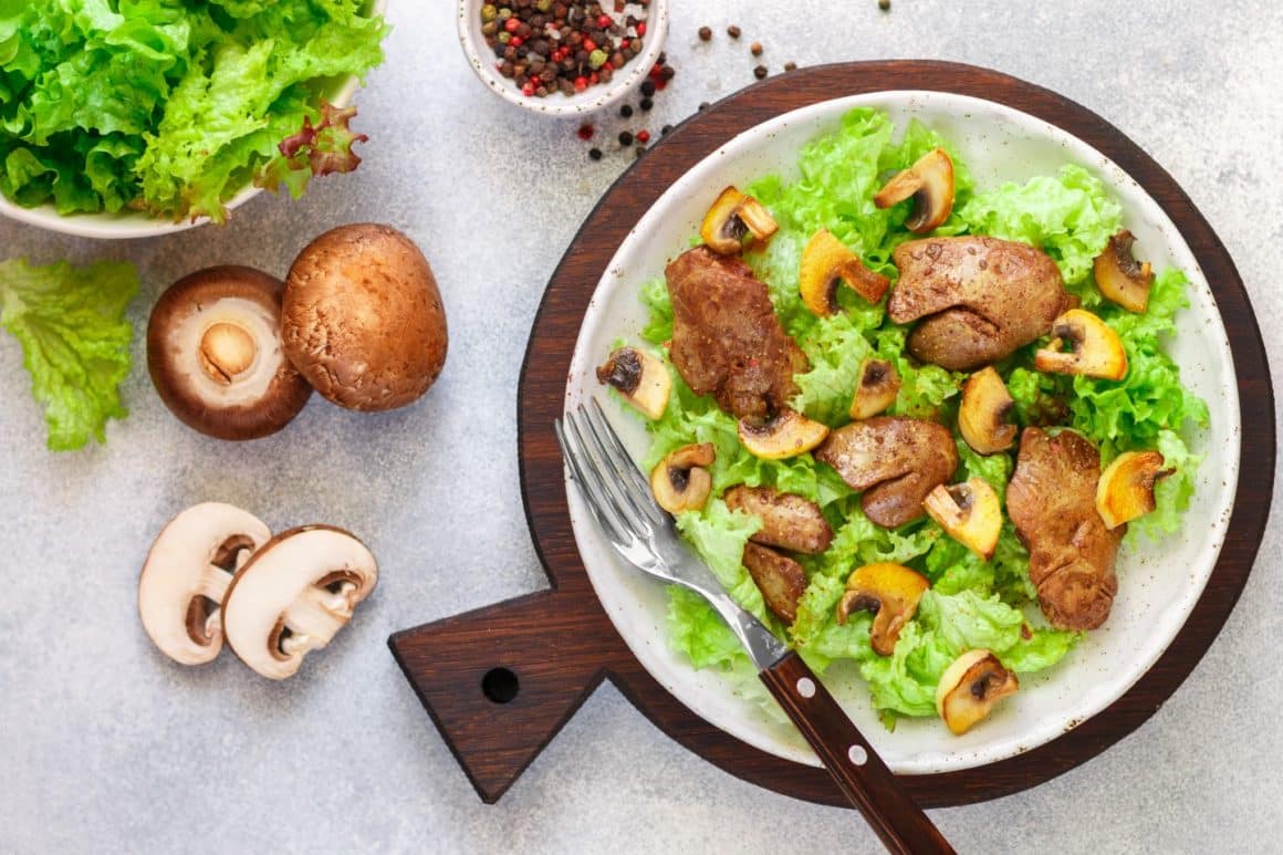 un bol cu mai multe ingrediente folosite la prepararea unei salate calde salate calde, așezat pe un tocător de bucătărie, rotund, alătur de câteva ciuperci și frunze de salată