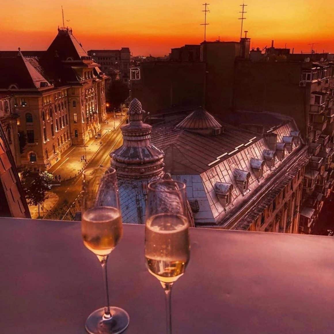 2 pahare de sampanie fotografiate pe balustrada terasei de la Cismigiu Bistro, iar in fundal priveșiste panoramica, de sus, asupra orașului la asfintit.  restaurante la înălțime din București