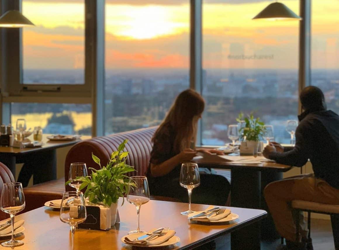 Cuplu care ia masa la restaurant in fata unor geamuri mari, la apus, cu vedere panoramica asupra orasuljui. restaurante la înălțime din București
