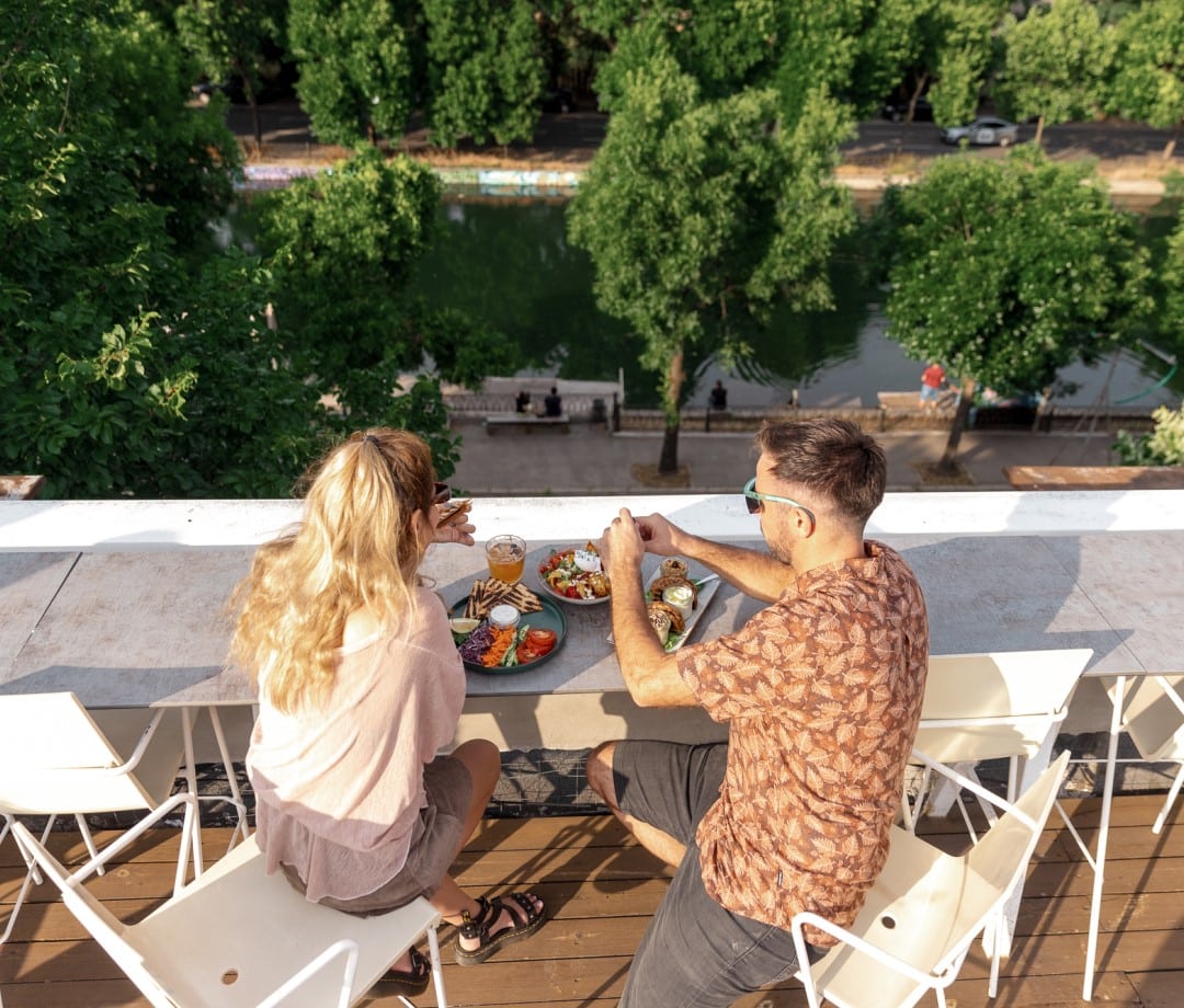 un cuplu ia masa pe terasa la Deschis gastrobar, situata la inaltime, cu privire spre raul Dambovita, vara, cu copacii inverziti. Restaurante la înălțime în București