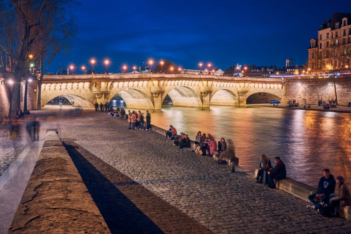 mai multe grupuri de oameni stau asezati pe bordura de pe aleea pietonala care merge de-a lungul raului Sena in Paris. Seara, cu un pont iluminat in fundal. 