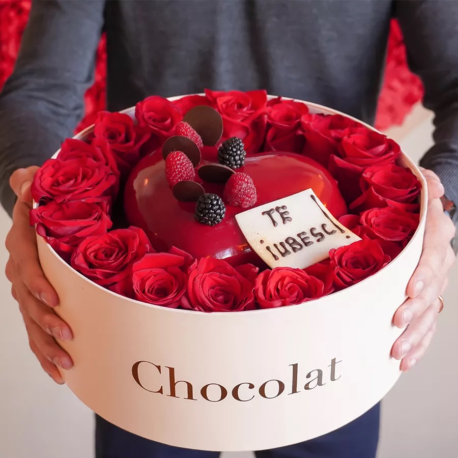 cutie de Valentines Day cu tort de bezea roz, in forma de inima, asezat printre trandafiri