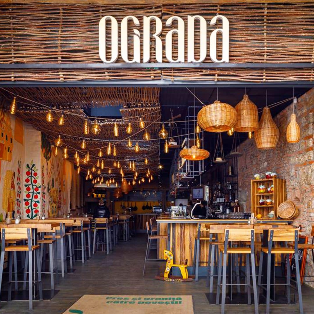 intrarea in restaurantul Ograda din Brașov, cu o roaba si ladite cu legume la intrare, si mese de partea cealalta, iar in fundal interior luminat cu multe siruri de becuri atarnate