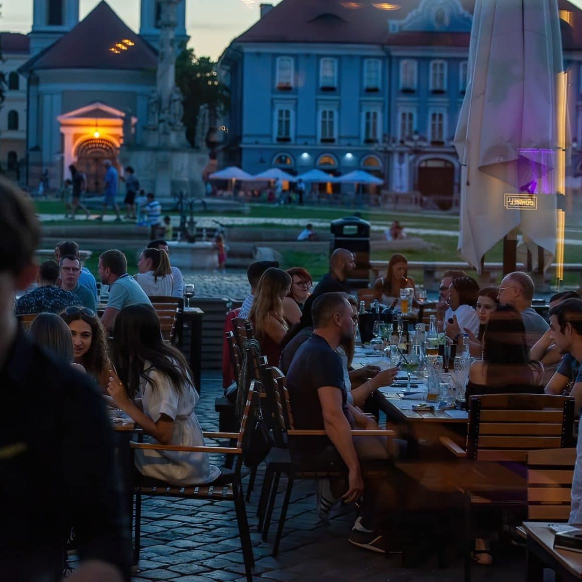 mai multi oameni iau masa la terasa, pe inserat, in Piata Unirii din Rimișoara
