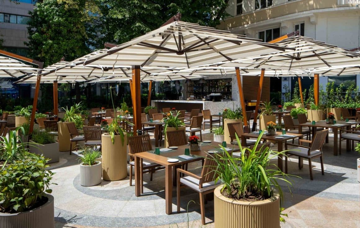 Terasa restaurantului Roberto's fotografiata ziua vara,, cu mobilier de lemn si umbrele mari, albe, multe plante decorative
