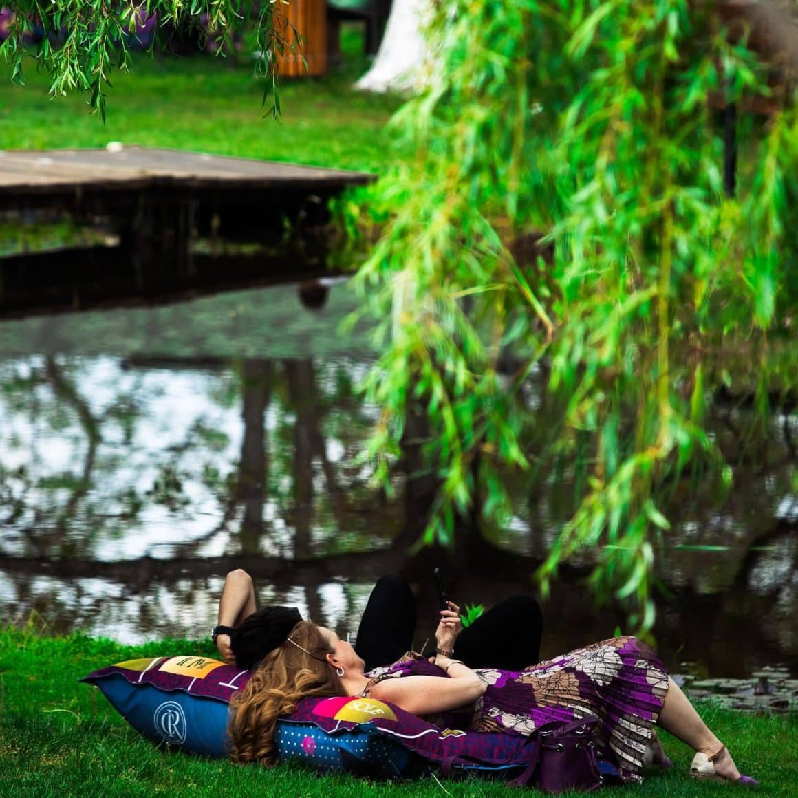 cuplu se relaxeaza culcati pe iarba pe malul unui lac la Acacia Village. Restort cu restaurante lângă București