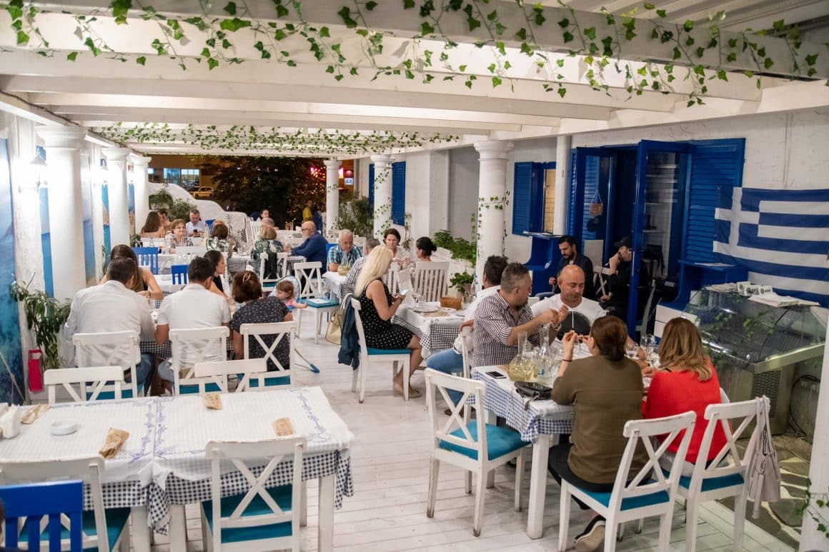 mai multi oameni iau masa la terasa restaurantului Santorini, unul din cele mai bune restaurante grecești din București. Terasa amenajata in stil grecesc, cu alb si albastru