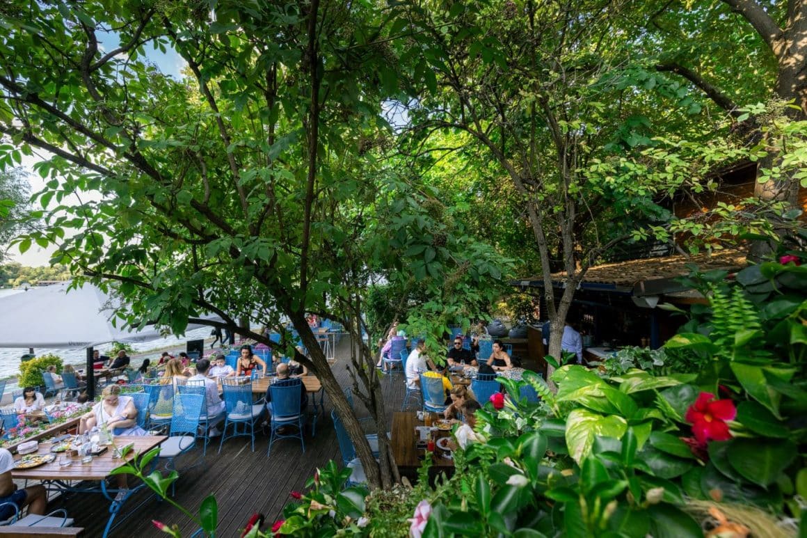 terasa restaurantului Pescaruis, fotografiata vara, cu mesele pline de oameni care iau masa la umbra copacilor deși.