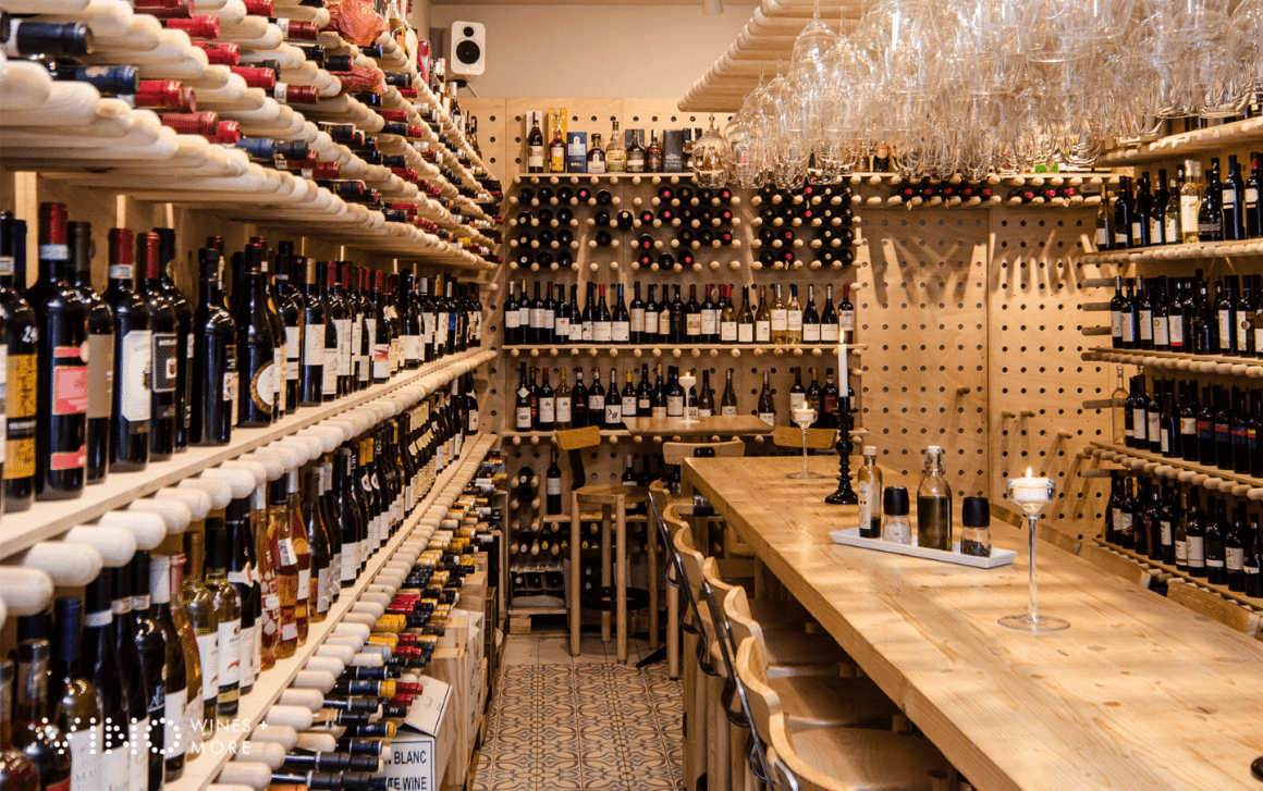 masa lunga asezata in dreptul unor pereti plini cu rafturi cu sticle de vin, la Vino wines & More. Wine Bars București